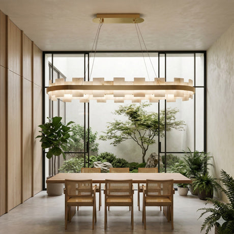 A modern dining room with a wooden table, six chairs, lush indoor plants, floor-to-ceiling windows overlooking a minimalist garden, and a striking Montyra LED Panelled Alabaster Linear Chandelier in soft white & brass.