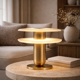 The Luxelle LED Double Tiered Alabaster Table Lamp in soft white and brass sits on a round stone table next to two closed books in a softly lit living room with neutral decor and a vase of dried flowers in the background.