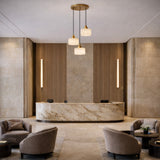 A modern hotel lobby with marble reception, beige stone walls, vertical wood paneling, Brinex 3 Light Cluster Alabaster Pendant Light in soft white and brushed brass, plus gray armchairs around dark tables for a sleek minimalist look.