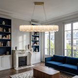 A stylish living room features the Valeris 14 Light Layard Panel Alabaster Linear Chandelier in soft white & brass, a marble fireplace, built-in bookshelves, navy blue sofa, large windows, and a patterned rug on herringbone wood floors.