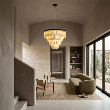 A modern, minimalist living room with neutral tones featuring the Auravel 9 Light Small Tiered Alabaster Chandelier in soft white and matte black, an olive green sofa, wooden chairs, a textured rug, built-in shelves, and floor-to-ceiling windows.