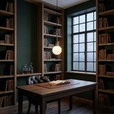 A cozy home library with tall bookshelves, a wooden desk, leather chair, and an Astra 1 Light Alabaster Single Pendant Light in Soft White & Brushed Brass glowing softly by the window on a cloudy day.