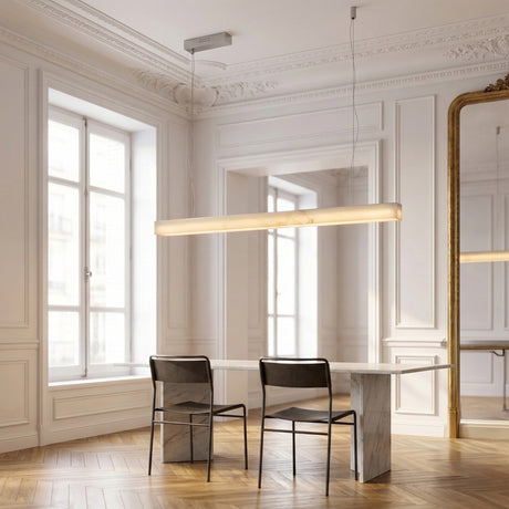 A minimalist dining room with two black chairs at a marble table, the Alba Large Alabaster Linear Suspension Light - Soft White above, large windows, white ornate walls, herringbone wooden floor, and a tall mirror reflecting the scene.