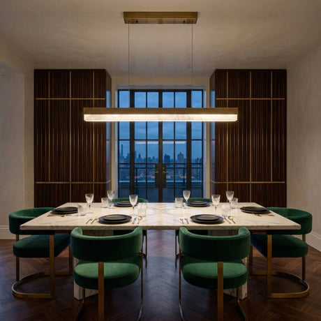A modern dining room features a marble table for six, green velvet chairs, the Linea LED Small Alabaster Linear Suspension Light in soft white & brushed brass overhead, dark wood paneling, and large windows revealing a city skyline at dusk.