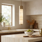 A modern kitchen featuring the Elviro LED Small Slim Tube Alabaster Single Pendant Light in soft white & brushed brass, marble countertops, a bowl of green apples, a gold faucet, and abundant natural light through the window.