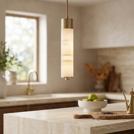 A modern kitchen featuring the Elviro LED Small Slim Tube Alabaster Single Pendant Light in soft white & brushed brass, marble countertops, a bowl of green apples, a gold faucet, and abundant natural light through the window.