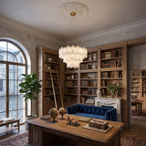 Elegant home library with tall bookshelves, a wooden desk with globes and a model building, blue velvet sofa, large window, potted plant, and the Caldrisa 16 Light 2 Tier Layer Panel Alabaster Chandelier in soft white & brass hanging above.