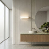 Minimalist bathroom with a large round mirror, the Velisse LED Horizontal Alabaster Wall Light in soft white, a floating vanity with ribbed wood front, round vessel sink, small bonsai tree, and a frosted glass window.