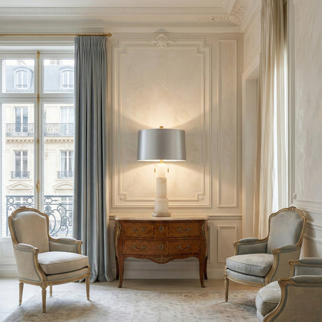 Elegant living room with ornate white walls, vintage wooden chest, Eloria 1 Light Stacked Alabaster Table Lamp in Brass & Soft White, two classic armchairs, tall curtains, and large windows overlooking Parisian-style buildings.