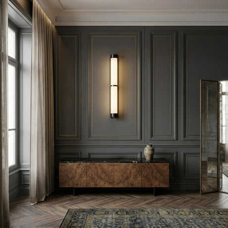 A modern, elegant room features dark paneled walls, tall curtains, a wooden sideboard with a marble top, a decorative vase, and a large mirror illuminated by the Arelis LED Linear Alabaster Wall Light – Soft White & Black above the sideboard.