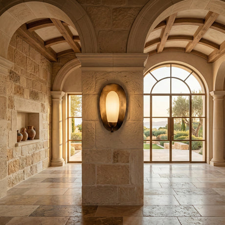 A Mediterranean-style hallway with stone walls, arched doorways, built-in nooks with pottery, large windows overlooking a garden, and the Orlisse LED Sculpted Alabaster Wall Light in soft white & brushed bronze as a feature sconce.