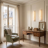 A bright, elegant room with a classic wooden desk, books, vases, a mirror, cushioned armchair and blanket, large windows with sheer curtains, and warm Aureline LED Small Sculpted Column Alabaster Wall Light on cream-paneled walls.