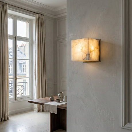 The Calmora 1 Light Block Alabaster Wall Light - Soft White, featuring a marble-like finish, is mounted on a light wall in an elegant room with tall windows, cream curtains, and a wooden table set for tea.