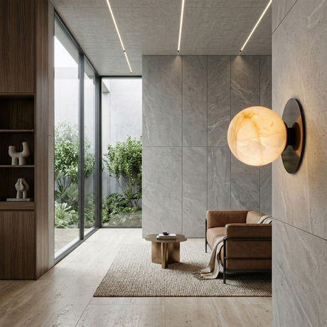 Modern living room with gray stone walls, a brown leather sofa, round wooden coffee table on a textured rug, large windows overlooking greenery, and the Noctyra 1 Light Alabaster Wall Light - Soft White & Black featured in the foreground.