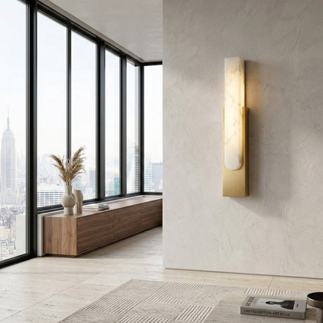 A modern, minimalist living room with floor-to-ceiling windows, wood console, light rug, neutral décor, a Nerava LED Large Slim Column Alabaster Wall Light in soft white and brass, and a city view with the Empire State Building.