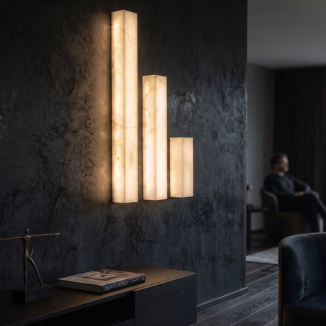 Valmera LED Slim Vertical Alabaster Wall Light in Small, shown alongside taller versions on a dark textured wall, glowing warm white in a modern living room setting.
