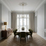 A modern dining room with a rectangular table, eight green velvet chairs, gold chandelier, large window with beige curtains, wooden sideboard featuring the Virella 1 Light Faceted Stone Table Lamp in Brass & Soft White, and herringbone wood floors.
