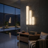 Valmera LED Slim Vertical Alabaster Wall Light in Small size, three staggered rectangular columns glowing warm white on a dark stone wall in a living room setting.