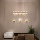 Petraia Linear Alabaster Chandelier in brushed brass, illuminated above matching wall sconces in a neutral living room with plant, sofa and alabaster side table.