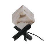 The Kalythra 1 Light Faceted Alabaster Table Lamp features a marble-textured, cube-shaped shade and intersecting matte black base. This sculptural piece includes a power cord extending from the base.