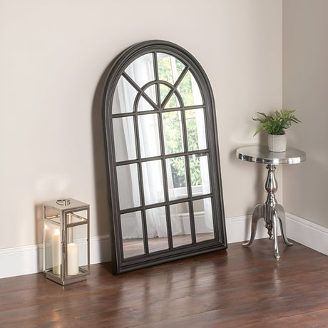 A Gothic elegance permeates the scene with the Arco Arched Leaner Mirror in black, measuring 119x80cm, leaning against a beige wall. On the left, a decorative lantern with candles casts warm glows, while on the right, a small silver table hosts a vibrant potted plant. The entire setup unfolds gracefully on a wooden floor.