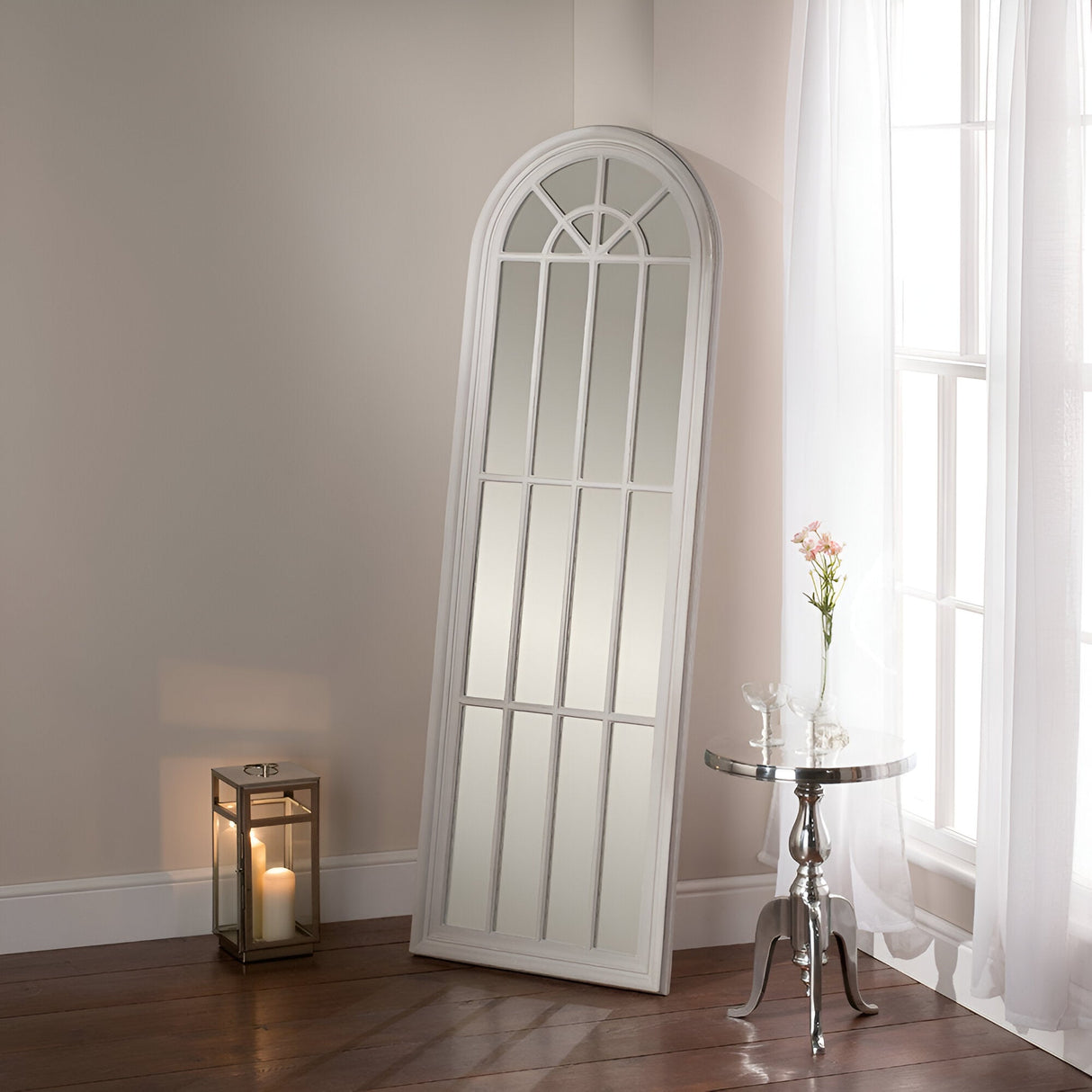 The Arco Arched Leaner Mirror Grey - 119x80cm, featuring a stunning white arched windowpane design and a grey wood frame, rests elegantly against a light-colored wall, offering a touch of modern elegance. A metal side table holds two glasses alongside a pink flower. On the floor nearby, a lantern with a lit candle casts a warm glow, while white curtains grace the adjacent window.