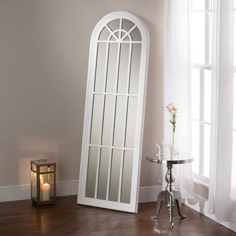 The Arco Arched Leaner Mirror in white, measuring 119x80cm, leans elegantly against a beige wall beside a large window, filling the room with natural light. Nearby, a small round table holds a glass vase of flowers and is complemented by a lantern with a lit candle on the wooden floor.