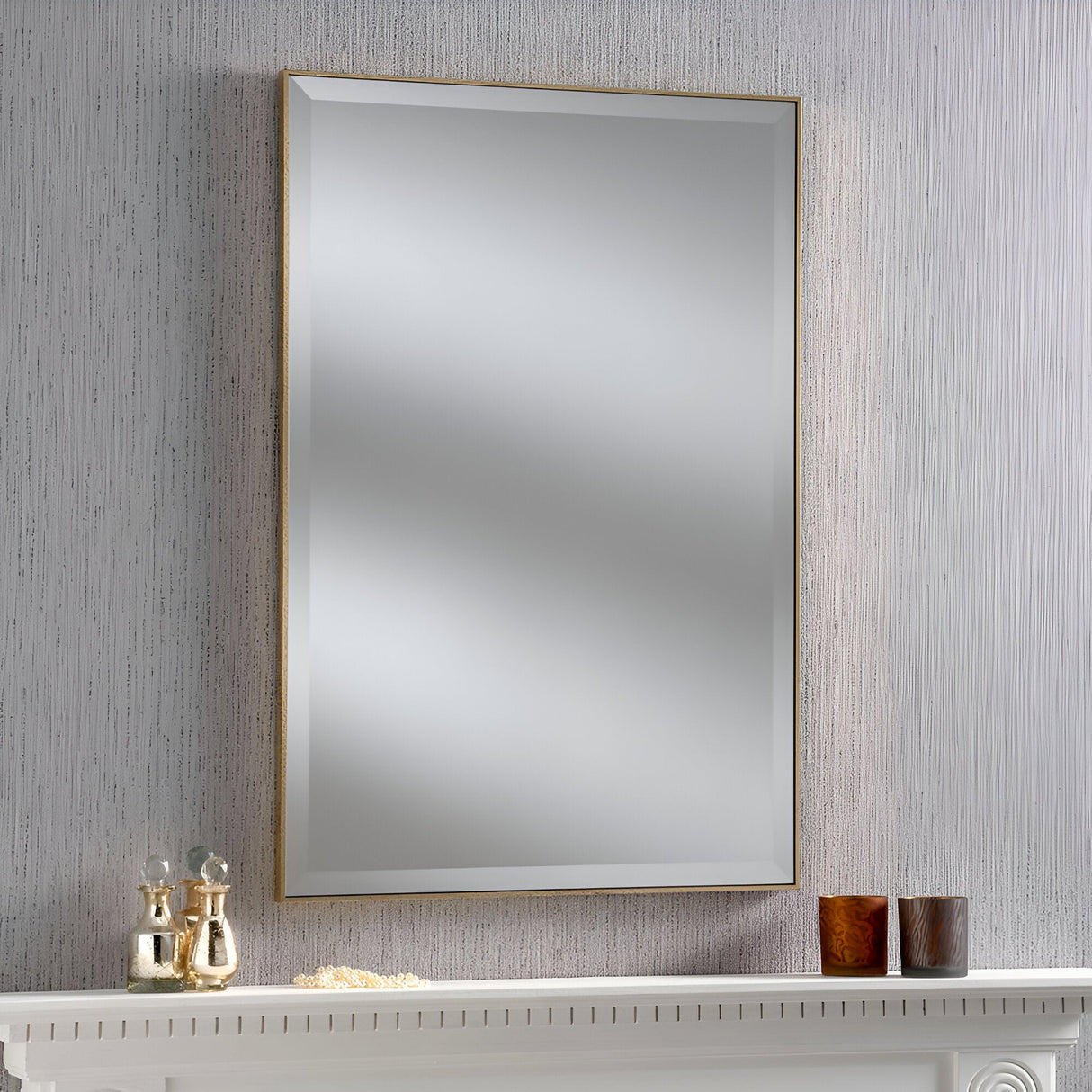 The Arden Rectangle Wall Mirror in gold, featuring a thin frame, hangs on a textured wall above a white mantel. The mantel is adorned with home decor, including two small vases and candles in brown holders, set against a subtle grain pattern.