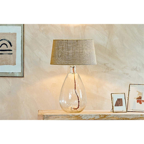 Baba Large Tall Recycled Glass Lamp Base in Clear, teardrop-shaped with a chrome neck collar, shown with a woven shade on a console table in a hallway setting.