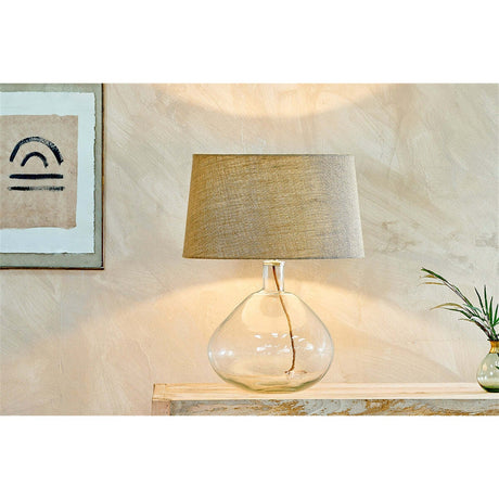 Baba Large Wide Recycled Glass Lamp Base in clear glass with a wide bulbous shape and linen drum shade, shown lit on a console table in a neutral-toned room.