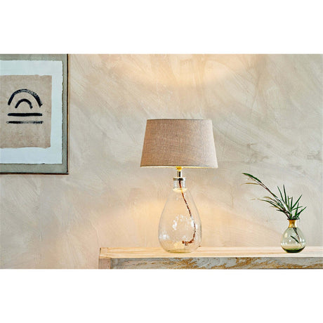 Baba Small Tall table lamp with teardrop-shaped clear recycled glass base and taupe fabric shade, shown lit on a wooden console table beside framed art and a small vase.