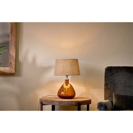 Baba Small Wide recycled glass table lamp in burnt amber with a tapered neutral shade, shown on a wooden side table beside an armchair in a living room setting.