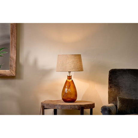 Baba Small Tall table lamp with a burnt amber recycled glass teardrop base and linen shade, shown lit on a wooden side table beside an armchair.