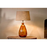 Baba Small Tall Recycled Glass Lamp Base in Burnt Amber, teardrop glass body with chrome neck collar, shown lit with a grey tapered shade on a wooden stool in a bedroom setting.