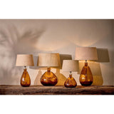 Baba Small Tall Recycled Glass Lamp Base in Burnt Amber, a narrow teardrop glass base with hammered facets, shown among similar lamps with linen shades on a wooden shelf.