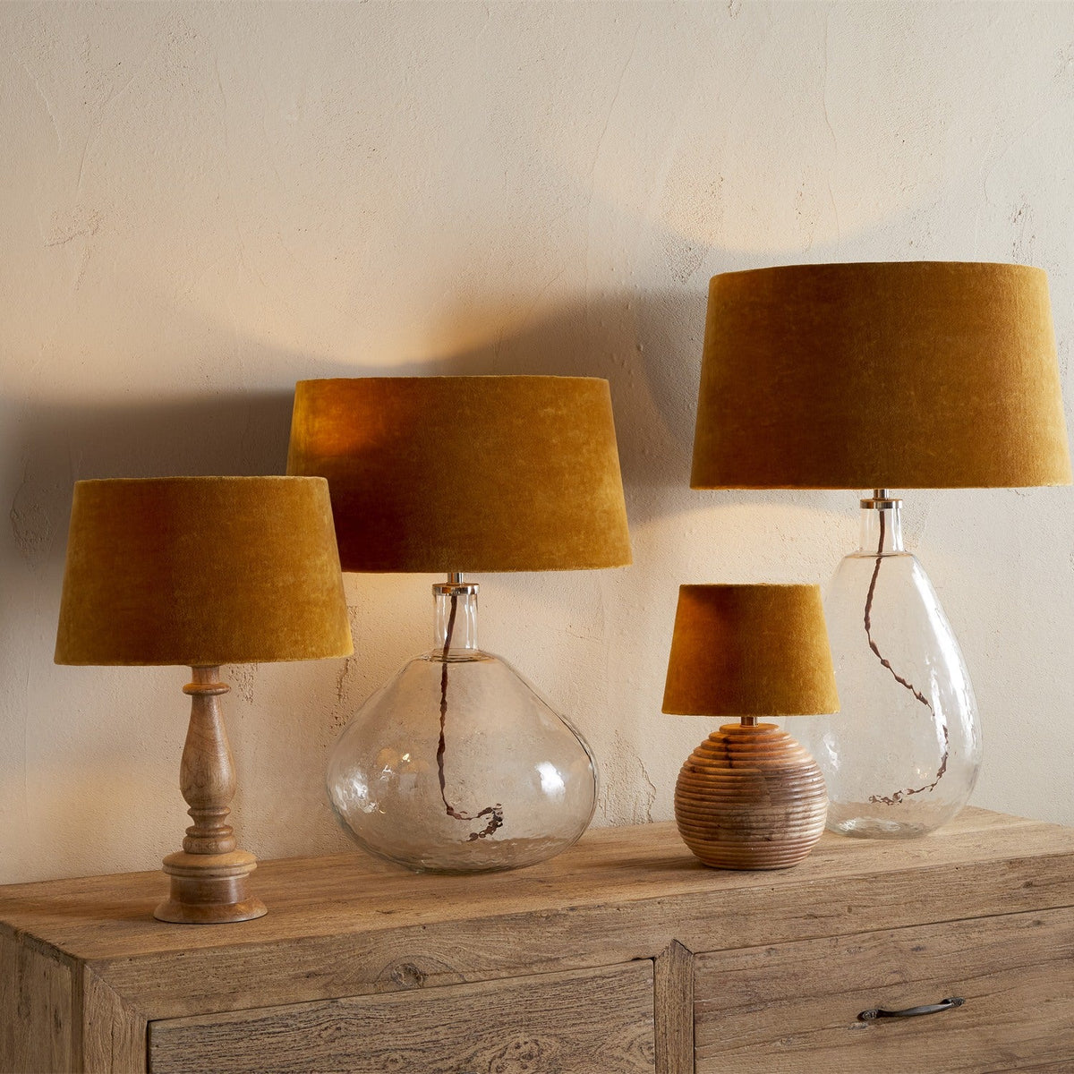 Group of table lamps with dark ochre velvet tapered drum lampshades on wood, ceramic, and glass bases, styled against a neutral wall atop a wooden sideboard.