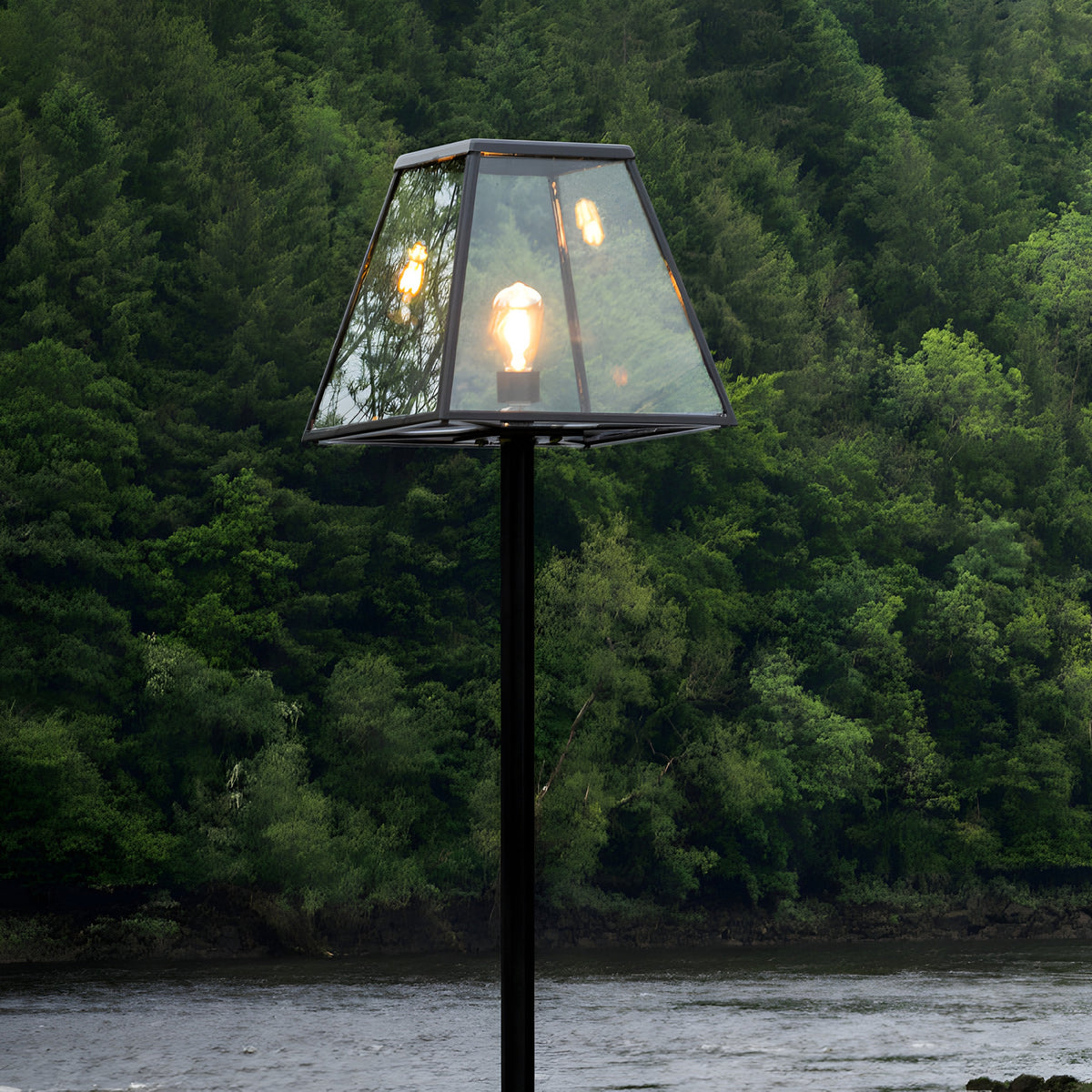 Contemporary black galvanised steel lamp post, 2.7m, with tapered glass lantern head and single exposed bulb, set against a wooded riverside backdrop.