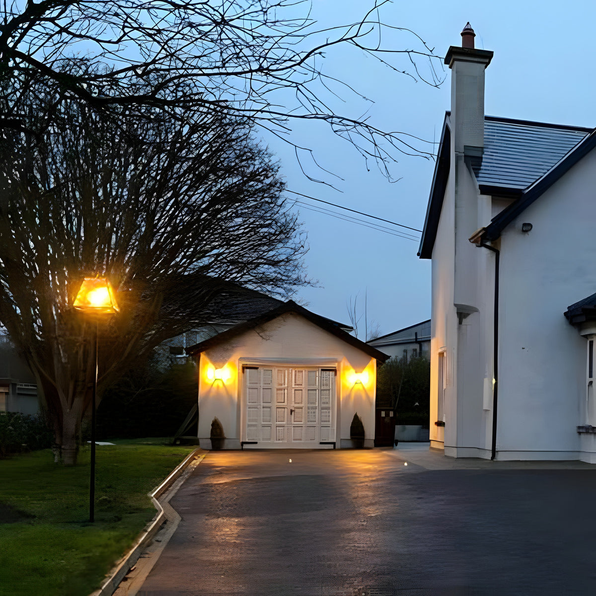 Contemporary black galvanised steel lamp post, 2.7m, with tapered lantern head and hand-cut glass panels, illuminated on a driveway beside a white house at dusk.