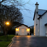 Contemporary black galvanised steel lamp post, 2.7m, with tapered lantern head and hand-cut glass panels, illuminated on a driveway beside a white house at dusk.