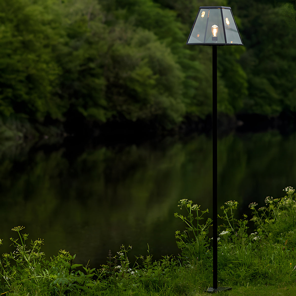 Contemporary Galvanised Steel Lamp Post 2.7m in black with tapered glass lantern shade and lit bulb, standing beside a garden lake edged with greenery.