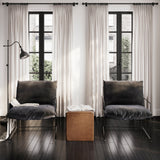 Pair of Brook accent chairs with black velvet seats and matt black metal frames, either side of a tan leather cube in a living room with tall curtained windows.