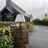 Large Contemporary Galvanised Steel Pillar Light in black, tapered glass-panelled design with exposed bulb, mounted on a stone gate pier outside a modern black-clad building.