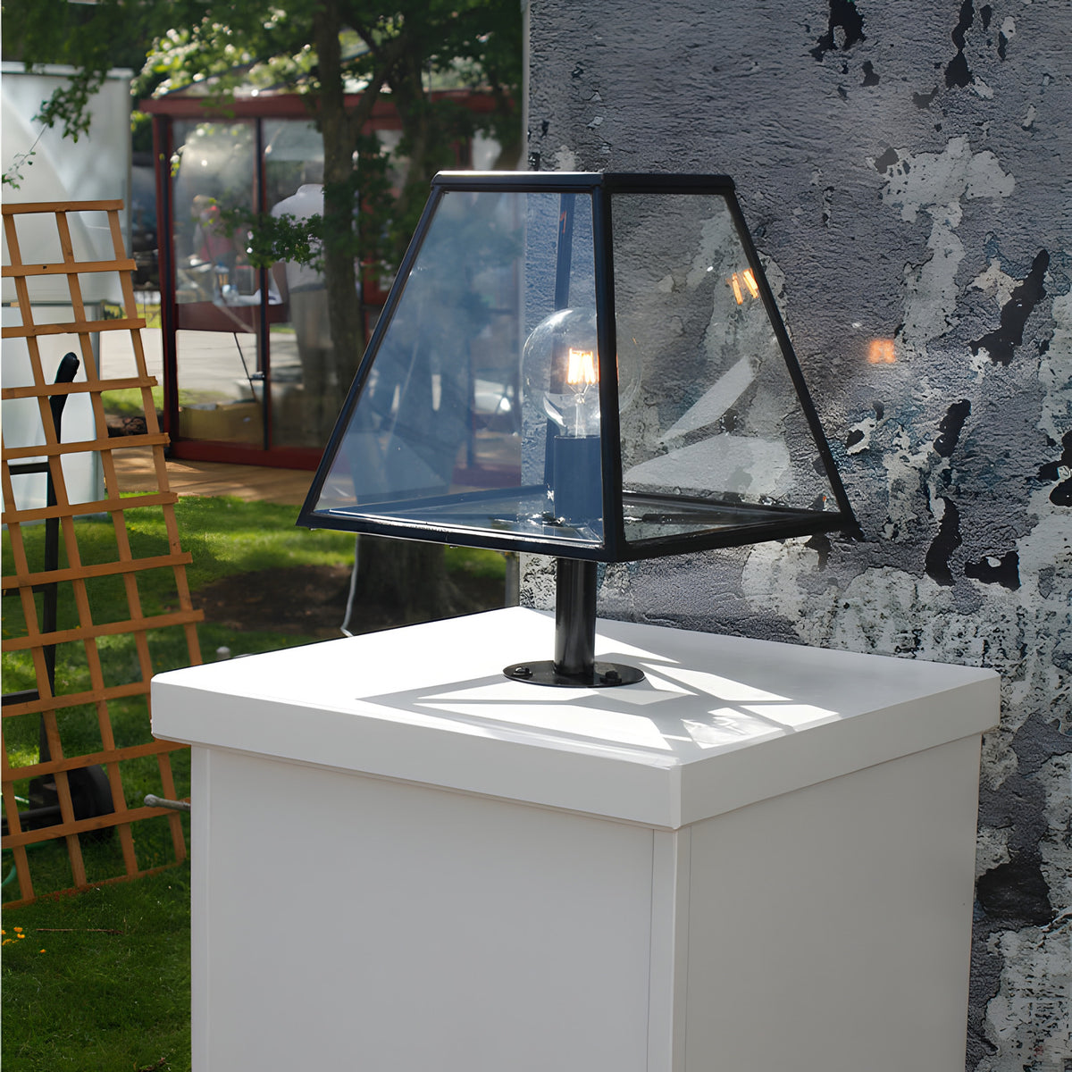 Large Contemporary Galvanised Steel Pillar Light in black, tapered glass panels with E27 bulb, shown outdoors mounted on a white plinth against a textured wall.