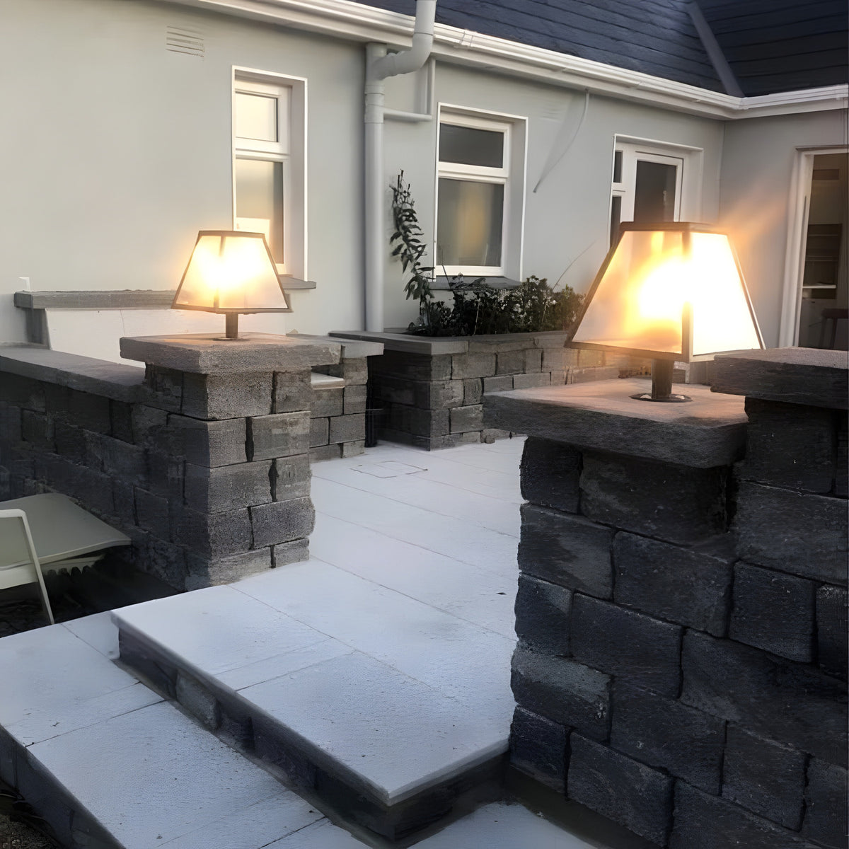 Small Contemporary Galvanised Steel Pillar Light in black, tapered glass shade illuminated atop stone gate piers along a garden patio wall.