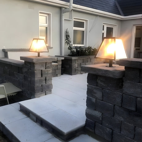 Small Contemporary Galvanised Steel Pillar Light in black, tapered glass shade illuminated atop stone gate piers along a garden patio wall.