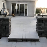 Small contemporary pillar light in black steel with tapered glass shade, mounted on a dark stone pier cap beside a patio and glass sliding door at dusk.