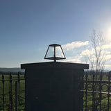 Small Contemporary Galvanised Steel Pillar Light in black, tapered glass shade atop a stone gate pier, set against a blue sky and countryside backdrop.