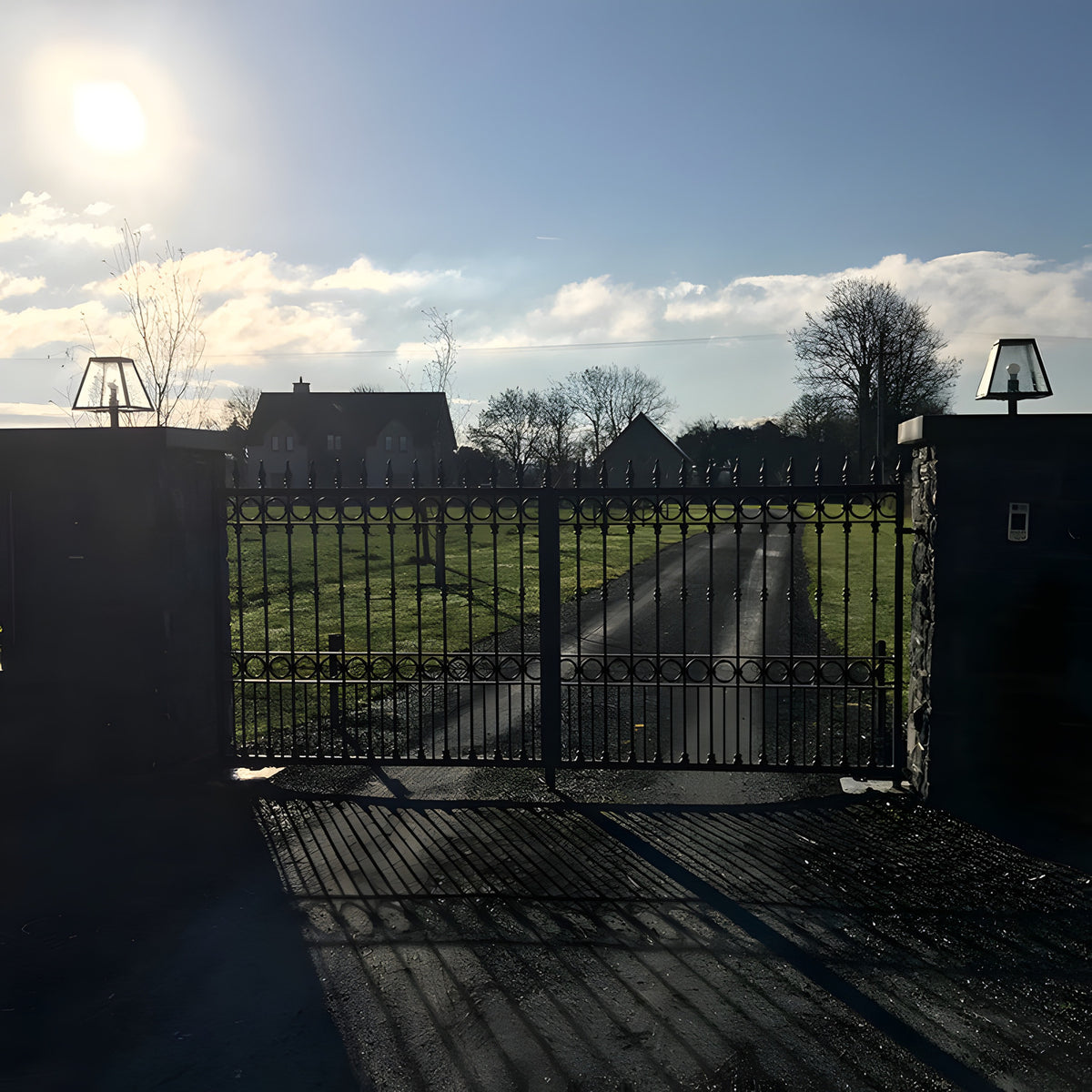 Small contemporary black pillar light mounted on a gate pier, tapered glass shade with pyramid top, installed at a wrought iron entrance gate.
