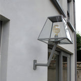 Large Contemporary Wall Lantern on an angled bracket in grey, mounted on an exterior wall beside a window, with a glass tapered shade and exposed globe bulb.