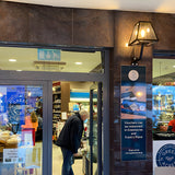 Small contemporary black outdoor wall lantern on an angled bracket, glass-panelled steel shade, shown mounted on a stone wall above a shopfront entrance.